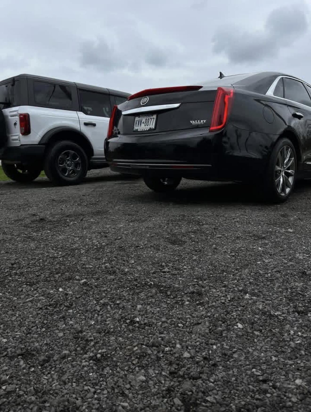 Black Cadillac sedan and white Ford Bronco parked on gravel under cloudy skies